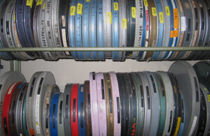 A number of movie canisters in a range of colours including silver, blue, red, pink and green. Many of them have been labeled in handwriting or with Dymo labeling tape. The canisters are housed on a rack.