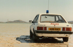 A Ford Escort is being used as a Police car. A blue flashing light has been affixed to the roof and the word 'Police' has been painted on the car boot.