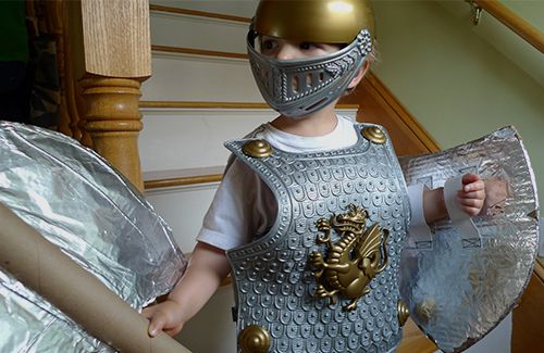 A child wearing a knight costume and holding a home-made shield and battle axe made out of cardboard and aluminium foil.